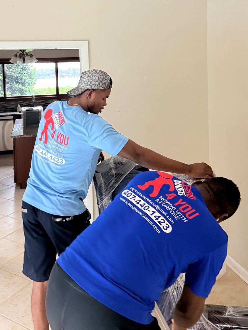 Two Legend Movers crew members in branded blue shirts stretch-wrapping furniture inside a home