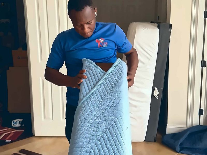 Legend Movers 4 You mover unfolding a quilted moving blanket in a bedroom to pad furniture