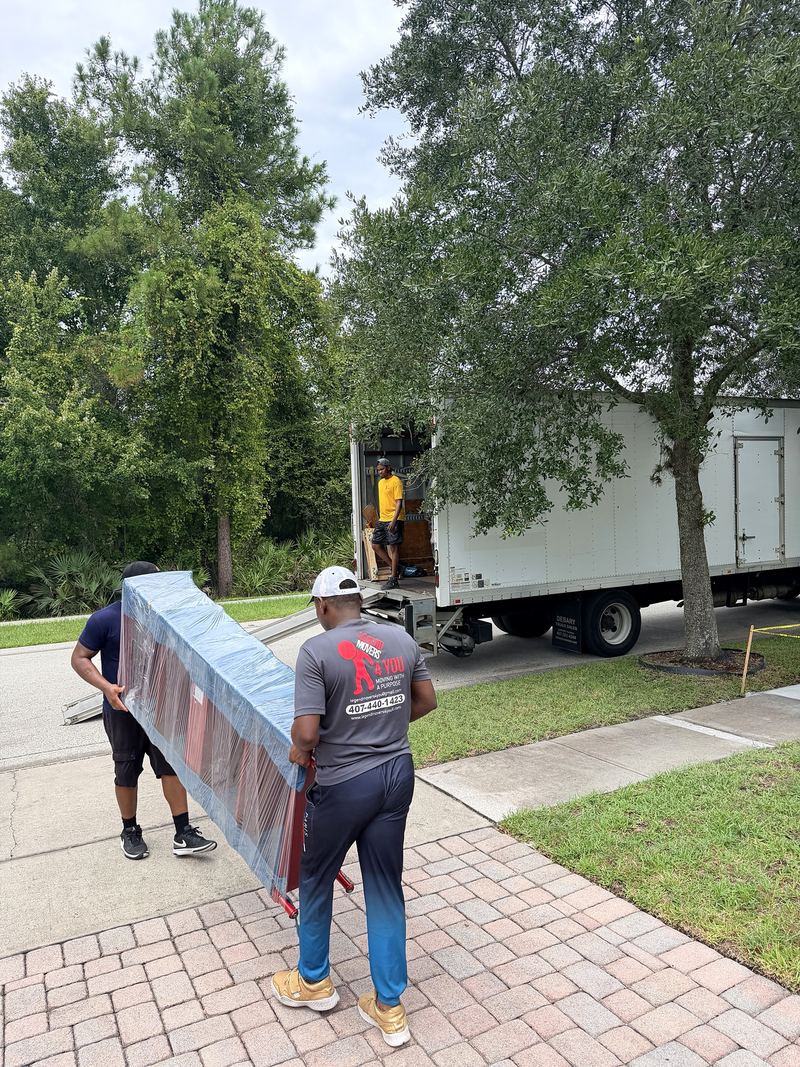Two movers carrying a wrapped piece down a front walkway to the truck