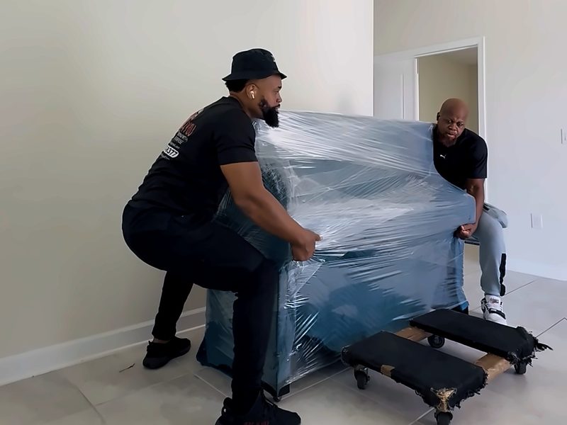 Two Legend Movers 4 You movers lifting a stretch-wrapped upright piano onto piano dollies